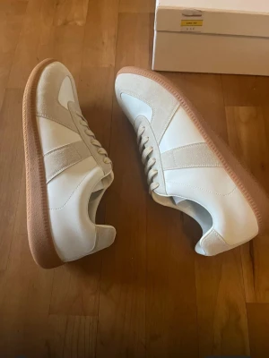 Maison Margiela gats - Stilrena vita Maison Margiela gats  med beige mockadetaljer och gummisula. Klassisk siluett med lågt skaft och snörning. Perfekta för en clean och trendig look. Materialmix av läder och mocka ger en lyxig känsla.