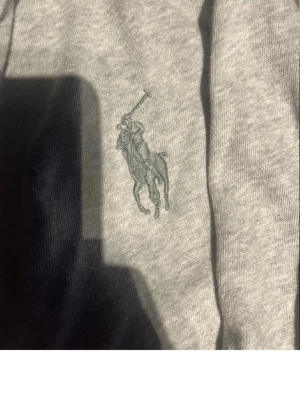 Grå zip hoodie från Polo Ralph Lauren - Klassisk grå zip hoodie från Polo Ralph Lauren med huva och vita dragsnören och tunnare tröja . Broderad logga i mörkgrått på bröstet och två stora fickor framtill. Tillverkad i mjuk bomullsblandning för en skön och avslappnad look.