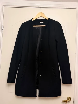 Svart klassisk kappa från Vero Moda - Stilren svart kappa från Vero Moda med rak passform och diskreta fickor framtill. Kappan har knäppning med tryckknappar och är helfodrad för extra komfort. Perfekt för dig som gillar en minimalistisk look.