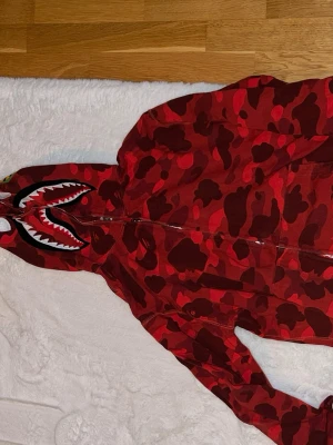 BAPE SHARK - Säljer en röd camo hoodie från BAPE med ikoniskt hajmotiv på huvan och tigerpatch på sidan. Hoodien har dragkedja hela vägen upp och är tillverkad i mjuk bomull. Perfekt för dig som vill sticka ut med streetwear-stil.