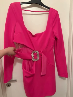 Rosa kort klänning med bälte - Snygg rosa kort klänning med lång ärm och markerad midja med ett brett bälte och glittrigt spänne. Klänningen har en unik cut-out detalj vid midjan och fyrkantig ringning. Perfekt för dig som vill sticka ut med en färgstark look.