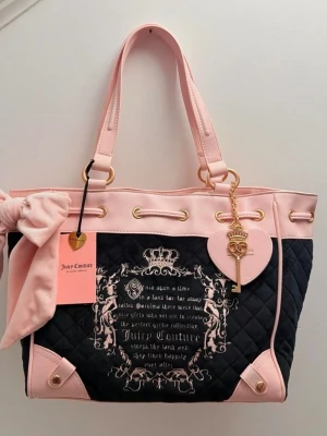 Juicy Couture  daydreamer fairytale tote  - Stilren quiltad handväska från Juicy Couture i svart och ljusrosa med dekorativ rosett, guldfärgade detaljer och nyckelberlock. Väskan har tryckt text och emblem på framsidan samt dubbla handtag och hjärtformad tag.