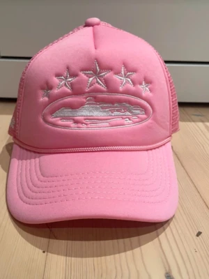 Rosa truckerkeps från Corteiz - Snygg rosa truckerkeps från Corteiz med vit brodyr framtill som visar stjärnor och ett motiv, samt broderad logga bak. Kepsen har mesh på baksidan och justerbar snapback. Perfekt för att lyfta din streetwear-look.