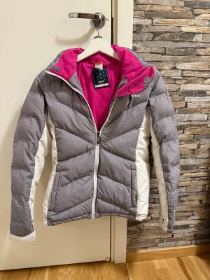 Grå och vit pufferjacka med rosa foder - Snygg pufferjacka från 8848 Altitude i grått och vitt med ett coolt rosa foder. Jackan har huva, dragkedja framtill och är vadderad för extra värme. Perfekt för kalla vinterdagar och har en sportig look med kontrastpaneler längs ärmarna.