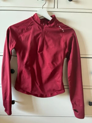 Röd långärmad träningströja GT Tech - Röd långärmad träningströja från GT Tech med halv dragkedja framtill och hög krage. Tröjan har tumhål i ärmsluten och är tillverkad i ett stretchigt funktionsmaterial som andas. Perfekt för löpning eller gym.