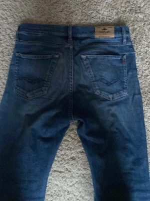 Blå raka jeans från Replay - Snygga blå jeans från Replay med raka ben och klassiska bakfickor med broderad detalj. Jeansen har en läderpatch med logga bak i midjan och är tillverkade i denim. Perfekta för en avslappnad och trendig stil.