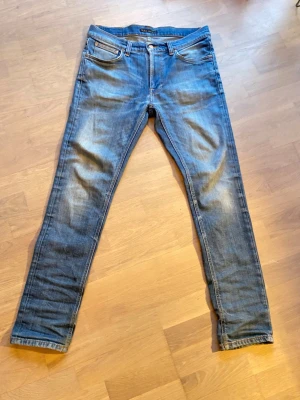 Blå slim fit jeans från Nudie Jeans - Säljer ett par blå slim fit jeans från Nudie Jeans med klassisk femficksmodell och snygga slitningar på låren och knäna. 