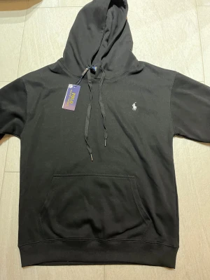 Polo Ralph Lauren Hoodie – Svart – Äkta – Storlek M - Riktigt fräsch Polo Ralph Lauren hoodie i svart, storlek M. Modellen är den klassiska med broderad pony-logo, känguruficka och dragsko i huvan. Sitter perfekt, clean passform, inga konstigheter.  ✔️ Äkta vara – QR-tag finns kvar (se bild) ✔️ Mjukt och tjockt bomullstyg ✔️ Inga defekter, inga fläckar, inga hål ✔️ Originaltaggen kvar – nästan som ny ✔️ Perfekt för både vardag och lager-på-lager outfits  Skick:10/10 Färg: Svart Storlek: M Passform: Normal  En hoodie som alltid går hem – tidlös, stilren 