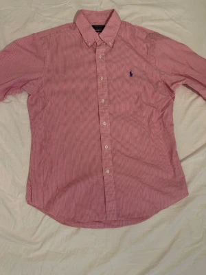 Rosavit randig skjorta Polo Ralph Lauren - Klassisk rosavit randig skjorta från Polo Ralph Lauren i slim fit-modell. Skjortan har långa ärmar, knappar framtill och den ikoniska blå logotypen broderad på bröstet. Tillverkad i mjuk bomull och perfekt för en stilren look.