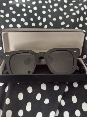 Svarta solglasögon All Cap One - Stilrena svarta solglasögon från All Cap One med markerad, fyrkantig båge och mörka glas. Kommer i ett snyggt fodral med logga. Perfekta för att ge din look edge och skydda ögonen mot solen.