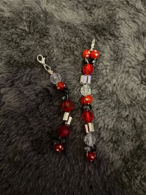 2 pack Berlocker med klolås  - Två handgjorda berlocker med pärlor i rött, svart och transparent, dekorerade med silvriga kubpärlor och klolås. Perfekt att fästa på väska, nyckelring eller som smyckesdetalj. Materialet är plast och metall.