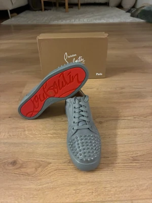 Grå Christian Louboutin sneakers - Snygga grå sneakers från Christian Louboutin med ikoniska röda sulor och nitar på tån. Skorna har lågt skaft, snörning och är tillverkade i läder. Perfekta för dig som vill sticka ut med en exklusiv och edgy stil. Priset går att diskutera 