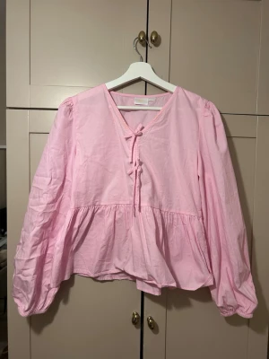 Ljusrosa blus med knyt framtill - Söt ljusrosa blus med v-ringning och dekorativa knytband framtill. Blusen har lång ärm med lätt puff och resår vid ärmslut samt en lös, flowy passform. Tillverkad i ett tunt, vävt material som känns luftigt och bekvämt.