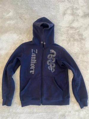 Rhinestone zip hoodie - Säljer en mörkblå hoodie med dragkedja från Zamioré. På framsidan finns ett stort tryck av en orm och texten 'Zamioré' i glittrande stenar. Hoodien har fickor framtill och en hög krage med luva. Perfekt för dig som gillar streetwear och unika detaljer.
