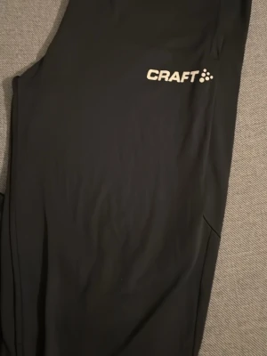 Svarta träningsbyxor från Craft - Snygga svarta träningsbyxor från Craft med logga på låret. Byxorna har en smal passform och är gjorda i ett stretchigt material som passar perfekt för träning och sportaktiviteter.