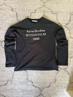 Acne Studios long sleeve  - Svart långärmad topp från Acne Studios med vit text 'Acne Studios STOCKHOLM 1996' på bröstet. Klassisk rund halsringning och rak passform. Perfekt för dig som gillar minimalistisk design och ikoniska märken.
