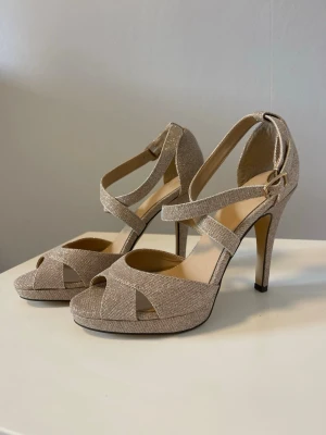 Glittriga beige pumps med höga klackar - Säljer ett par eleganta pumps i beige med glittrigt tyg och höga klackar. Skorna har öppna tår, platåsula och dubbla remmar med spänne runt ankeln. Perfekta för att lyfta din outfit med lite extra glamour.