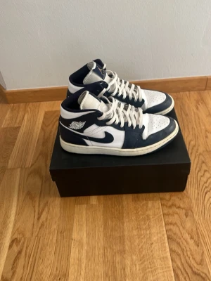 Nike Air Jordan 1 Mid Navy/Vit - Snygga Nike Air Jordan 1 Mid sneakers i marinblått och vitt läder. Klassisk siluett med högt skaft, perforerad tåbox och ikoniska Jordan Wings-loggan på sidan. Skorna har vita skosnören och en bekväm gummisula. Perfekta för dig som gillar streetwear och basketstil.