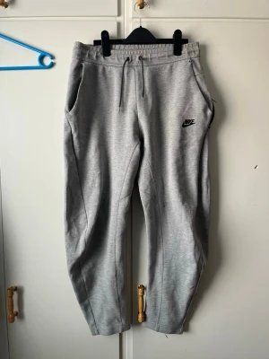 Gråa mjukisbyxor från Nike - Snygga grå mjukisbyxor från Nike med dragsko i midjan och sidofickor. Byxorna har en avslappnad passform och är tillverkade i mjukt material. Nike-logga broderad på vänster lår. Perfekta för chill eller träning.