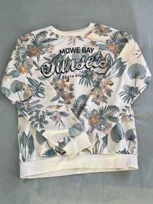 Krämvit sweatshirt med tropiskt tryck - Det är storlek 146/152 10-12år men den passar mig som är 14år och är 167cm lång. Krämvit sweatshirt med tropiskt blommönster och papegojor i grönt, beige och rosa. Texten 'Mowe Bay Sunsets South Atlantic' framtill. Rund halsringning och ribbade muddar. Perfekt för en avslappnad stil.