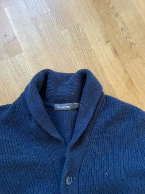 Mörkblå kofta från Dressmann - Mysig mörkblå kofta från Dressmann med sjalkrage och knäppning framtill. Koftan har två framfickor och är ribbstickad för en klassisk look. Perfekt att slänga på sig över en t-shirt eller skjorta för en avslappnad stil.