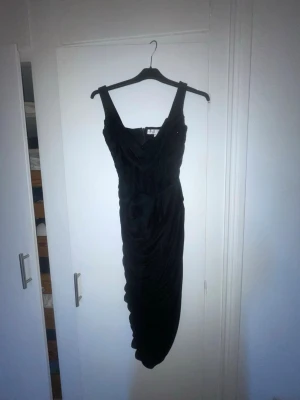 Svart långklänning scrunch  - Snygg svart långklänning med breda axelband och draperad front. Klänningen har en figurnära passform och är tillverkad i ett mjukt tyg som ger en elegant siluett. Perfekt för dig som vill ha en klassisk och stilren look.