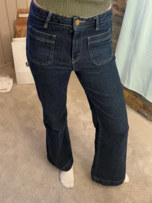 Mörkblå utsvängda jeans med fickor framtill  - Hej!🌸Jag säljer ett par snygga mörkblå jeans med bootcut passform och stora framfickor. Jeansen har hög midja, synliga sömmar och stängs med knapp och dragkedja. Perfekta för en avslappnad och trendig look.                         Modell: Front pocket flare jeans. Storleken 40, men passar dig som vanligtvis använd Medium. Väldigt fina!❤️