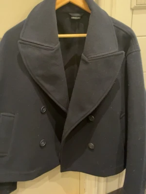 Marinblå skepparkavaj Gina Tricot XS - Stilren marinblå skepparkavaj från Gina Tricot med dubbelknäppning och bred krage. Jackan har två fickor framtill och är tillverkad i ett mjukt, ull-liknande material. Perfekt för dig som vill ha en klassisk look med modern touch.