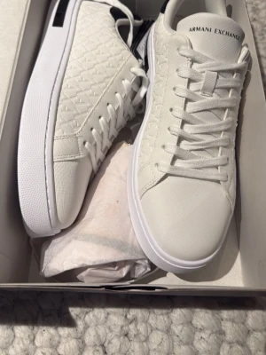 Vita sneakers från Armani Exchange - Stilrena vita sneakers från Armani Exchange med diskret mönstrad textil på sidorna, vita snören och svart detalj på hälen. Klassisk låg modell med rund tå och vit sula. Perfekta för dig som gillar en clean och modern look.