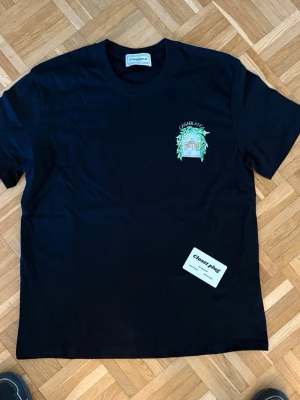 Svart Casablanca t-shirt  - Snygg svart t-shirt från Casablanca med färgglatt tryck på bröstet som visar en tennisscen omgiven av gröna blad och duvor. T-shirten har rund hals och korta ärmar, perfekt för en chill och stilren look.