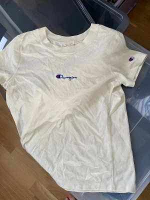 Beige Champion t-shirt i bomull - Snygg beige t-shirt från Champion i mjuk bomull. Klassisk passform med korta ärmar och rund halsringning. Champion-logga broderad på bröstet och ärmen. Perfekt för en avslappnad och sportig stil.