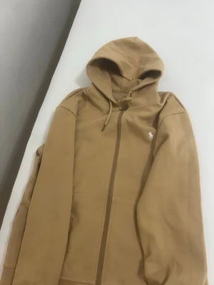 Beige hoodie från Polo Ralph Lauren - Snygg beige hoodie från Polo Ralph Lauren med dragkedja framtill och klassisk huva med dragsko. Ikoniska vita Polo-loggan broderad på bröstet. Tillverkad i mjukt bomullsmaterial och har två fickor framtill. Perfekt för en chill och stilren look.