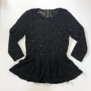 Setsblus med peplum och dragkedja - Svart topp i spets med peplumvolang nedtill och rund halsringning. Tre kvarts ärm och dekorativ guldig dragkedja i ryggen. Figurnära i modellen. 2010-tal stil. 