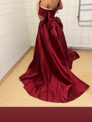 Vinröd långklänning med rosett - Elegant vinröd långklänning i satin med offshoulder-skärning och stor rosett i ryggen. Klänningen har en vacker, vid kjol och markerad midja, perfekt för bal eller fest. Materialet ger en lyxig känsla och klänningen har en klassisk, tidlös look.