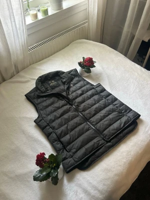 Svart dunväst från Morris - Snygg svart dunväst från Morris med quiltad design och hög krage. Västen har dragkedja framtill och två sidofickor. Perfekt för lager-på-lager och ger en stilren look. Klassisk Morris-logga på bröstet. Materialet är lätt och håller värmen bra.