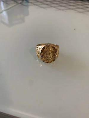 Guld ring - Unik guldfärgad ring med ett detaljerat lejonemblem på ovansidan och graverade mönster längs sidorna. Ringen har en klassisk, robust form och känns riktigt maffig på fingret. Perfekt för dig som gillar statement-smycken med kunglig känsla.