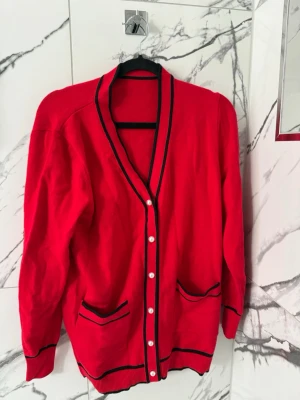 Röd kofta med svarta detaljer - Stilren röd kofta med svarta kanter och vita knappar framtill. Två fickor med svarta detaljer och ribbade muddar vid ärmslut och nederkant. Inga skador eller defekter. 