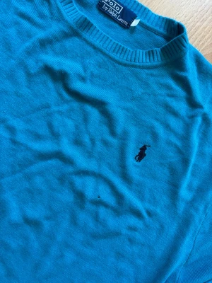 Blå tröja från Polo Ralph Lauren - Snygg blå tröja från Polo Ralph Lauren med klassisk rund halsringning och den ikoniska broderade loggan på bröstet. Tröjan har långa ärmar och ribbade muddar vid ärmslut och nederkant. Perfekt för en clean och stilren look. Har dock ett litet hål på framsidan. Axelmått = 53cm Längd = 66cm