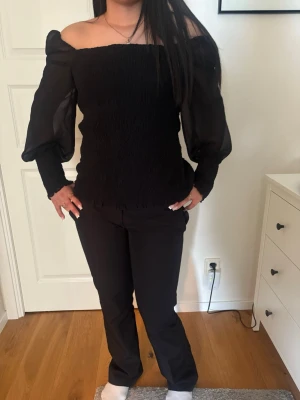 Svart offshoulder topp med puffärm - Trendig svart offshoulder topp med långa puffärmar i transparent tyg. Smockad överdel som sitter tight och markerar axlarna. Perfekt för dig som vill ha en stilren och snygg look med lite edge.
