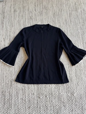 Svart topp med plisserade ärmar - Snygg svart topp med plisserade trekvartsärmar och vit kant längst ut på ärmarna. Toppens material känns mjukt och stretchigt, perfekt för en trendig look. Passformen är normal och toppen har en klassisk rund halsringning.