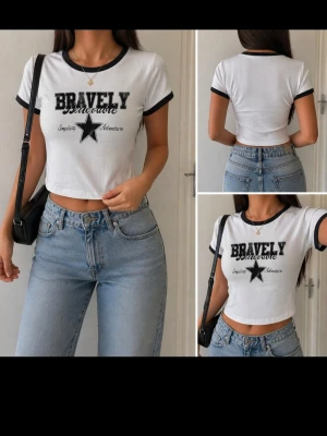 Vit croppad t-shirt med svart tryck - Snygg vit croppad t-shirt med svarta detaljer och texten 'BRAVELY' framtill. Svart stjärna och kontrastfärgade kanter vid ärmar och hals. Perfekt till jeans eller kjol för en cool och avslappnad look.