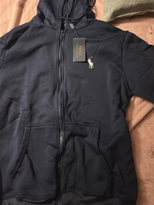 Mörkblå hoodie från Polo Ralph Lauren - Storlek L. Mörkblå hoodie med dragkedja från Polo Ralph Lauren. Klassisk design med huva, fickor framtill och den ikoniska vita loggan broderad på bröstet. Perfekt för en avslappnad och stilren look. Mjuk bomullsblandning för extra komfort.