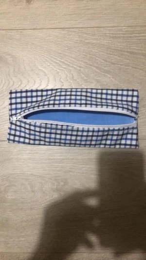 Rutigt pennfodral med dragkedja - Ett avlångt pennfack i tyg med blått och vitt rutmönster. Fodrad insida i ljusblått och vit dragkedja längs ovansidan. Perfekt för att förvara pennor eller småprylar. Mått: ca 9 cm brett. Enkel och stilren design utan synligt märke.