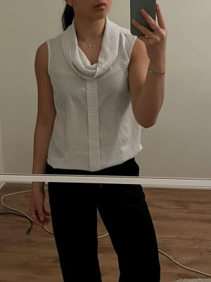 Polkadot blus - Snygg vit blus med polkadot och draperad krage. Ärmlös modell i lätt och luftigt material. Passar till både jeans och kostymbyxor. Liten mörkblå fläck på blusen (se bild 4). Vet ej exakta storleken, men skulle gissa storlek 38. Passar som S till M