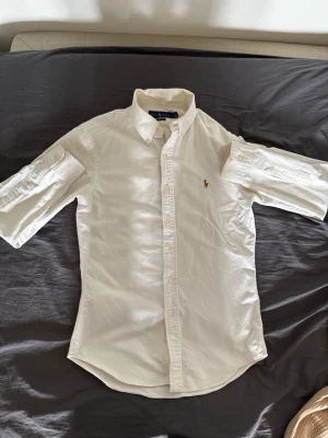 Vit skjorta från Ralph Lauren - Klassisk vit skjorta från Ralph Lauren med knappslå och button-down krage. Skjortan har korta ärmar som kan vikas upp och fästas med knapp, samt den ikoniska broderade loggan på bröstet. Perfekt till många olika stilar. Nypris 1100kr
