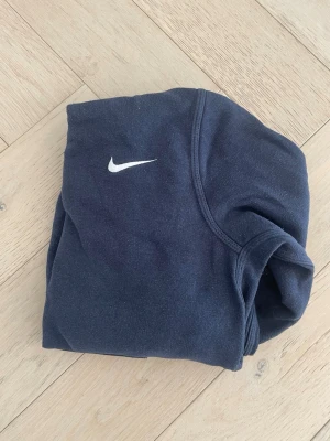 Mörkblå Nike hoodie med logga - En stilren mörkblå hoodie från Nike med klassisk vit swoosh-logga på bröstet. Hoodien har huva och en stor magficka framtill. Perfekt för en avslappnad och sportig look. Materialet känns mjukt och bekvämt.