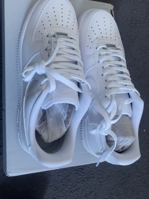 Nike Air Force 1 vita sneakers - Klassiska helvita Nike Air Force 1 sneakers med lågt skaft. Skorna har perforerad tåbox för ventilation, robust sula och ikonisk Swoosh på sidan. Tillverkade i läder med textildetaljer och vita skosnören. Perfekta för en clean och stilren look.