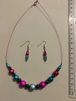 Färgglatt pärlhalsband med örhängen - Snyggt halsband med pärlor i metallicblått, rosa och svart på tunn rosa tråd. Matchande örhängen med blå och rosa droppformade pärlor ingår. Perfekt för att ge din outfit en färgklick och unik stil. Nickelfria