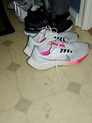 Nike Löparskor Air Zoom Pegasus  - Snygga vita Nike sneakers med grå swoosh och rosa detaljer på sulan. Ovandelen är i meshmaterial för bra andningsförmåga och skorna har svarta ränder på snörningen. Perfekta för en sportig och trendig look.