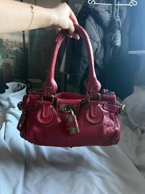 Röd Chloé Paddington handväska - Cool röd Chloé Paddington handväska i äkta läder med mässingsfärgade metalldetaljer, ikoniskt hänglås och nyckel. Väskan har dubbla handtag, dekorativa sömmar och nitar. Perfekt för dig som vill sticka ut med en statementväska.
